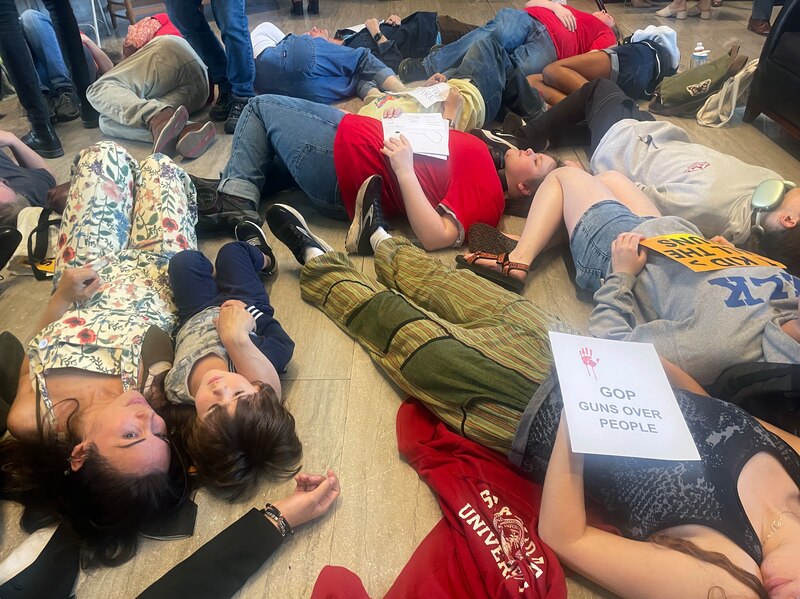 A group of people lie on the floor next to each other in a "die in" protest. Some are holding signs.