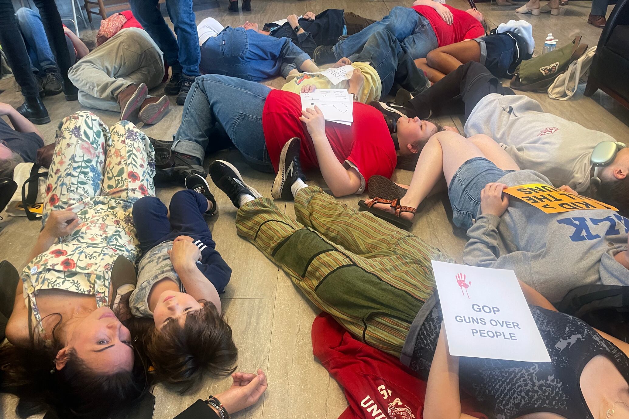 A group of people lie on the floor next to each other in a "die in" protest. Some are holding signs.