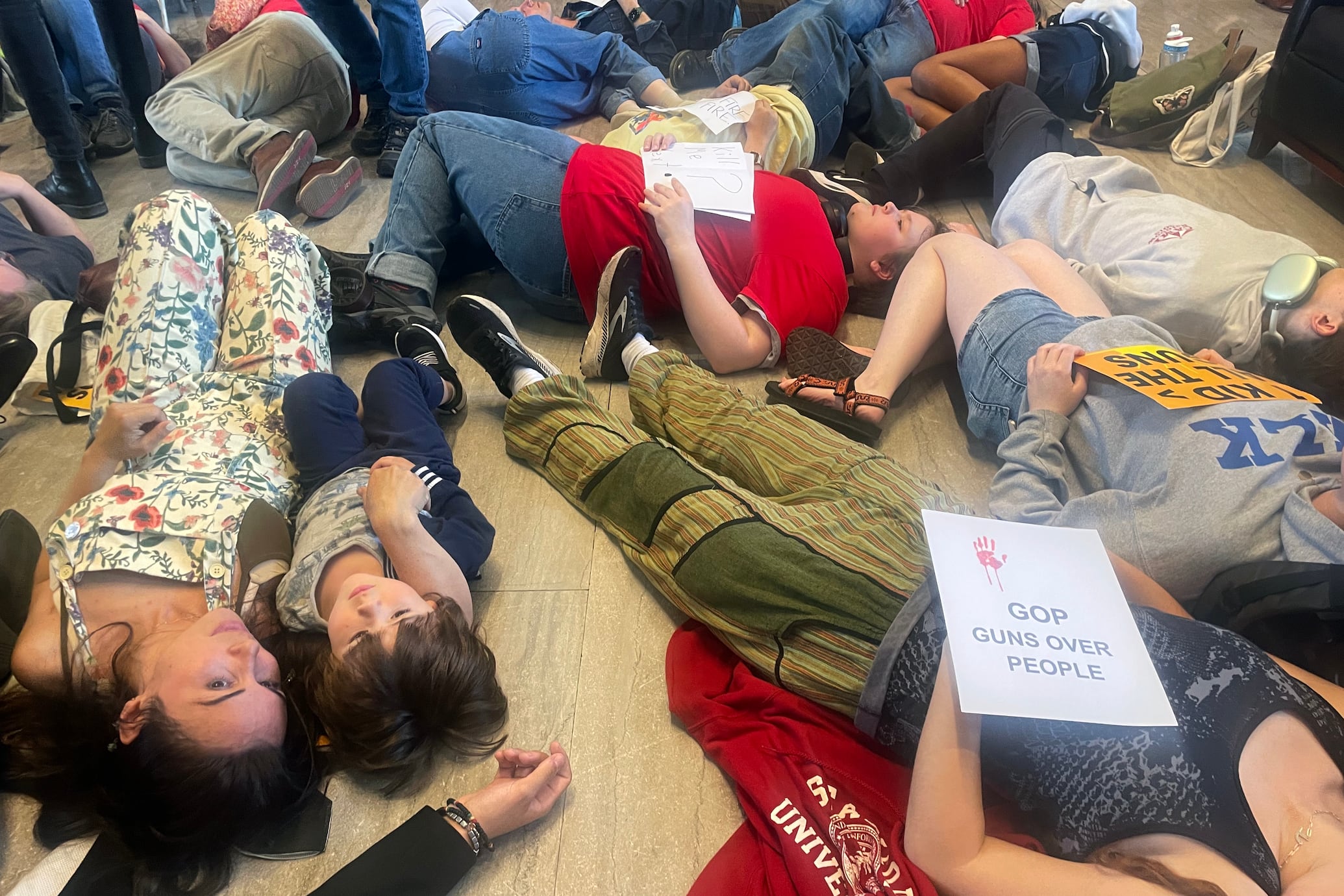 A group of people lie on the floor next to each other in a "die in" protest. Some are holding signs.