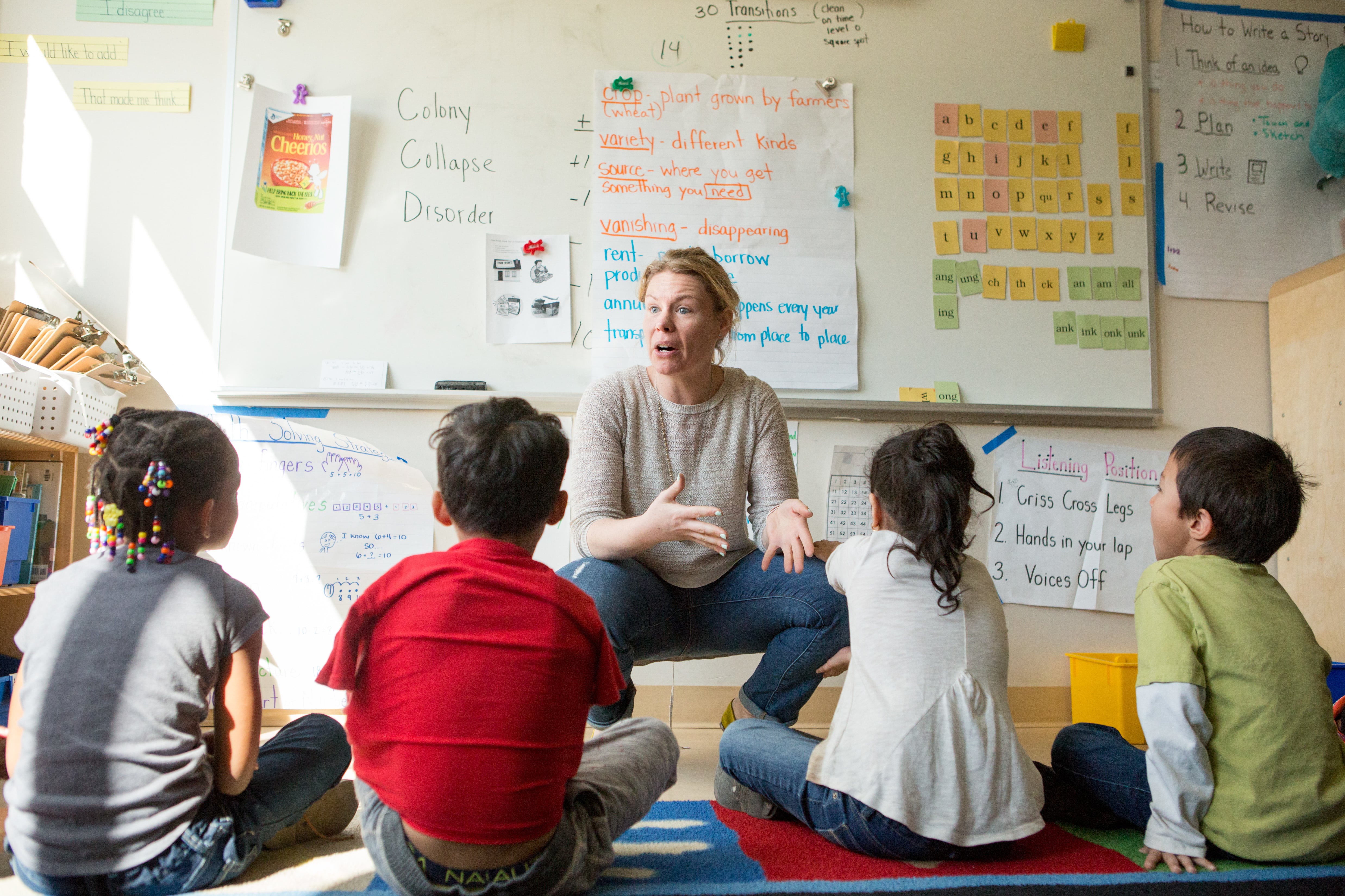 A woman in a white shirt and jeans crouches in front of and speaks to students sitting on the floor in a classroom. 