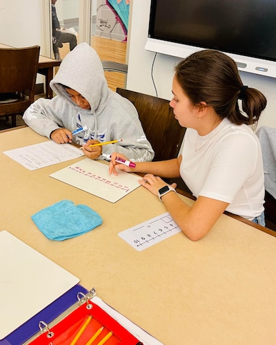 A female tutor wearing a white T-shirt sits at a table with a young student wearing a hoodie as they work together on a math problem.