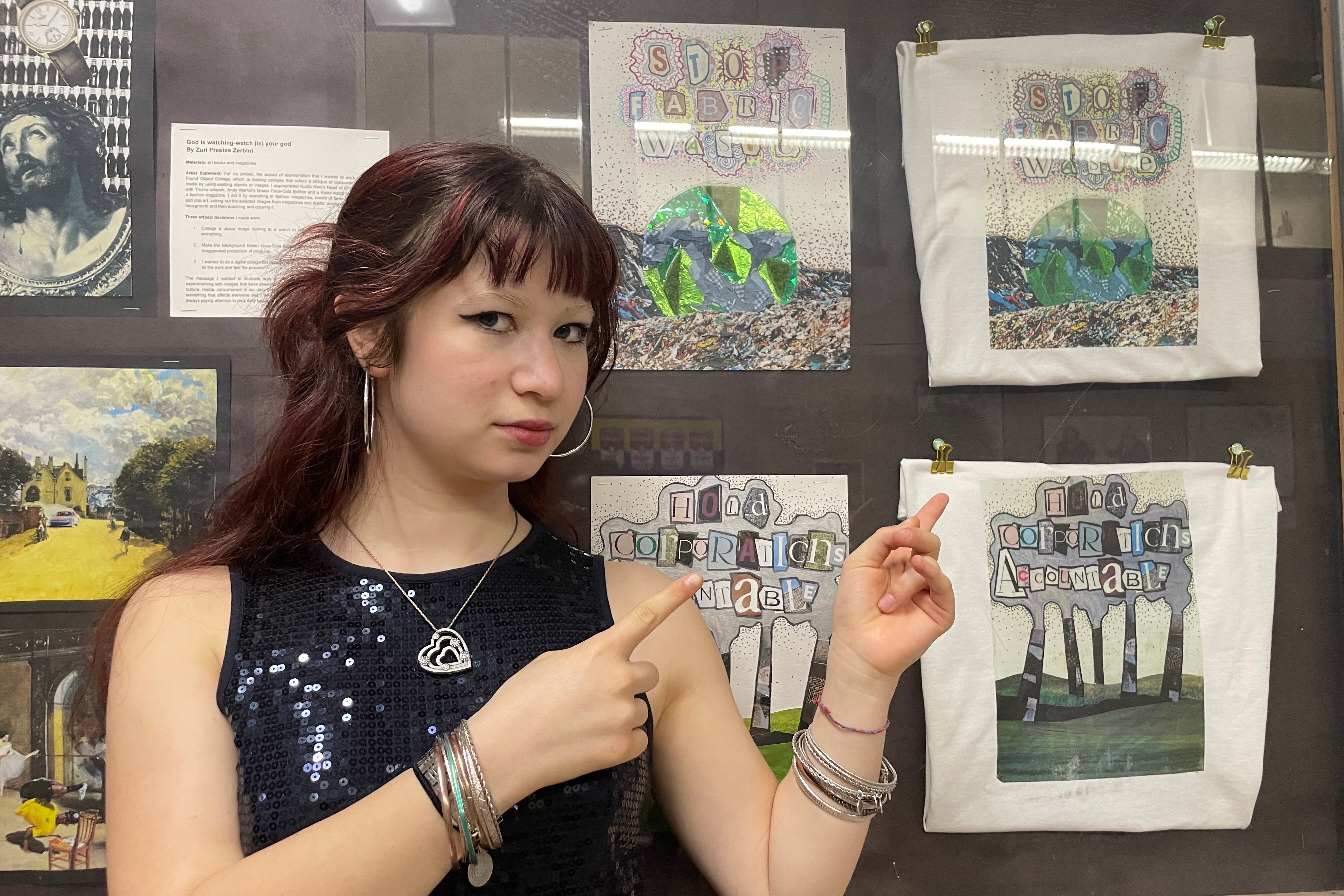 A high school student with long dark hair points to some drawings while posing for a photograph.