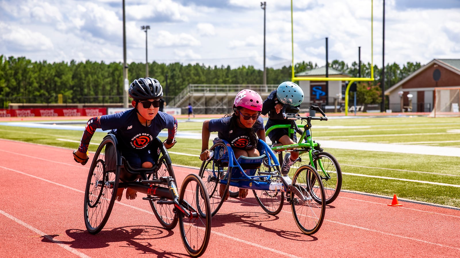 Three tweens, wearing helmets, in three-wheeled racing wheelchair on a track.