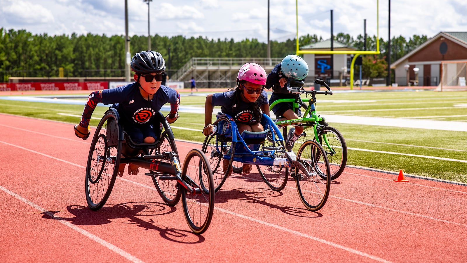 Three tweens, wearing helmets, in three-wheeled racing wheelchair on a track.