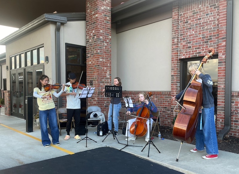 A photograph of a group of young people playing instruments outside of a large building.