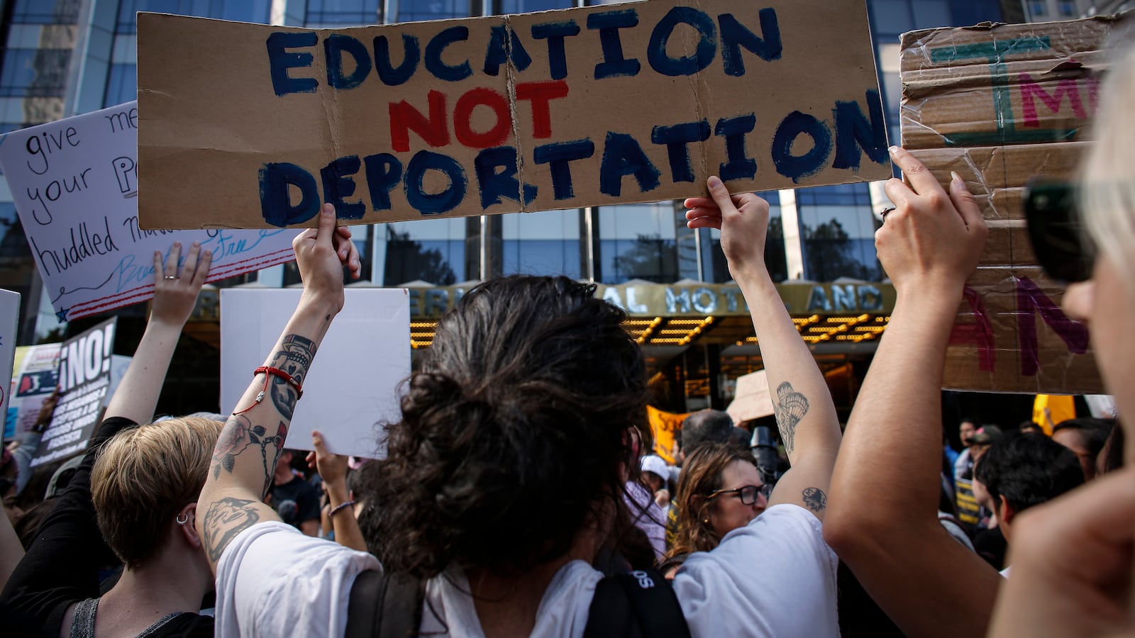 A group of people with their backs facing toward the camera hold up signs, one of which says, "Education Not Deportation."
