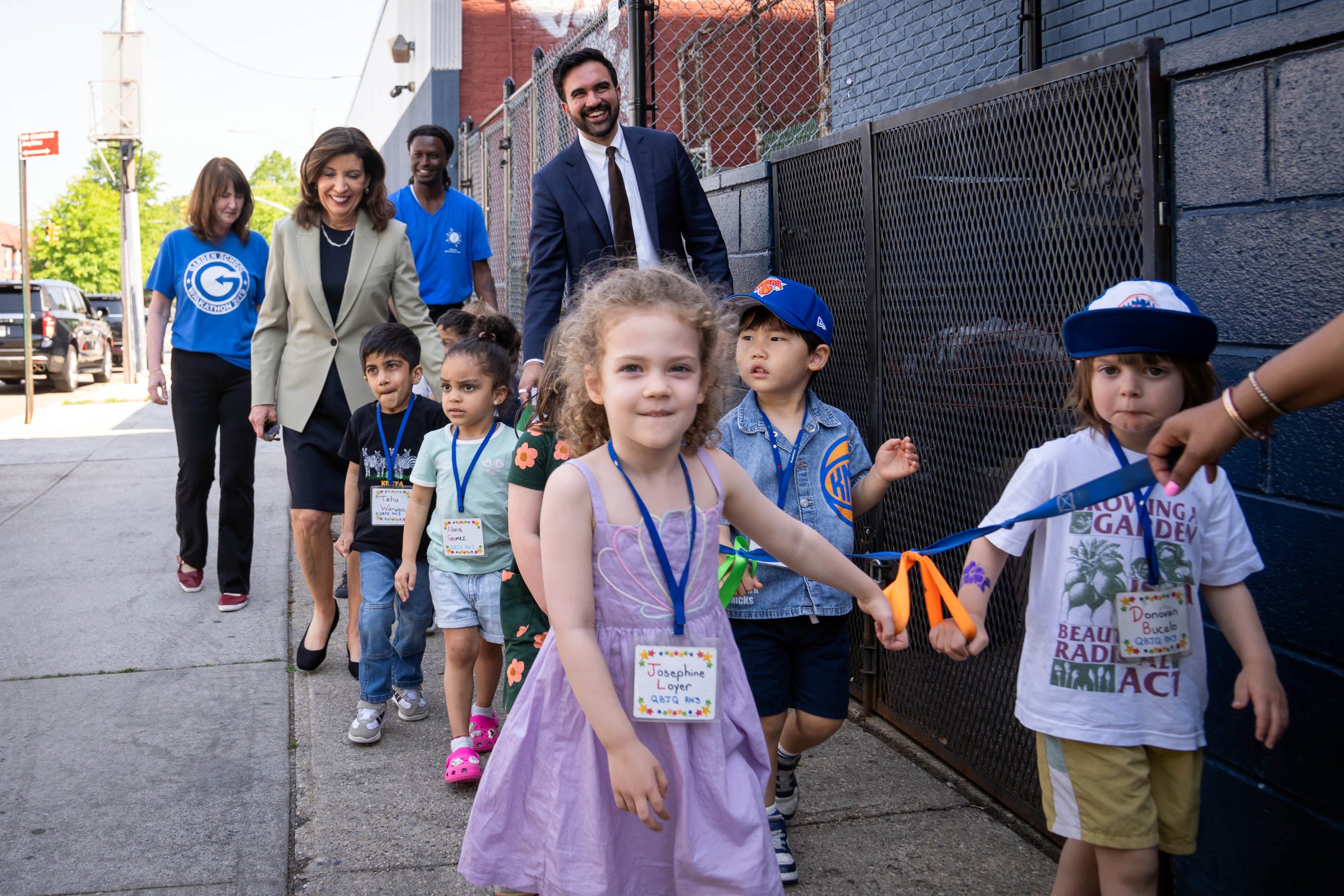 Adults walk behind a line of young students walking on a sidewalk outside.