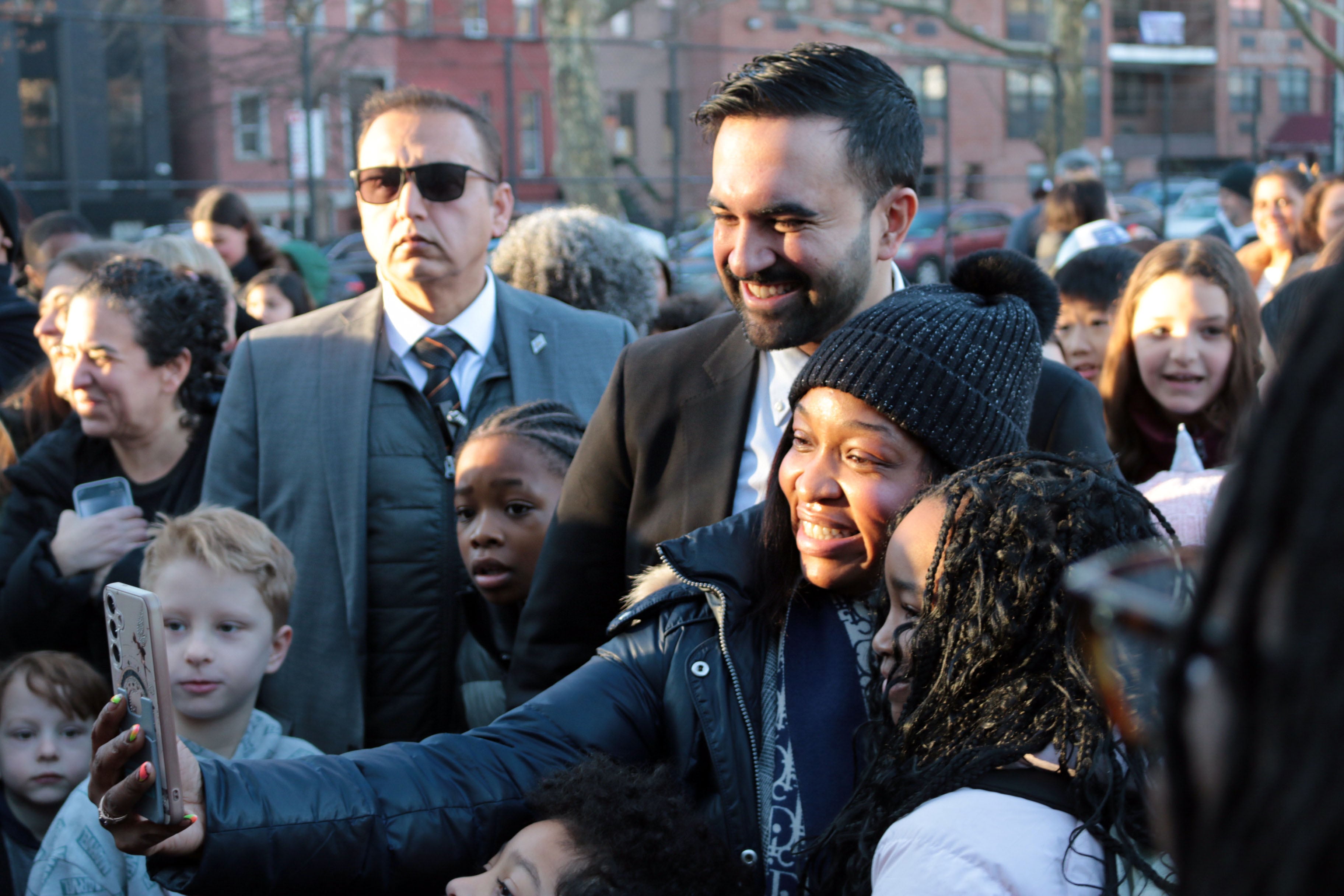 A photograph of a Black woman in a coat and hat holding out her cellphone to make a selfie with a man in a suit with a security agent and lots of students in the background.
