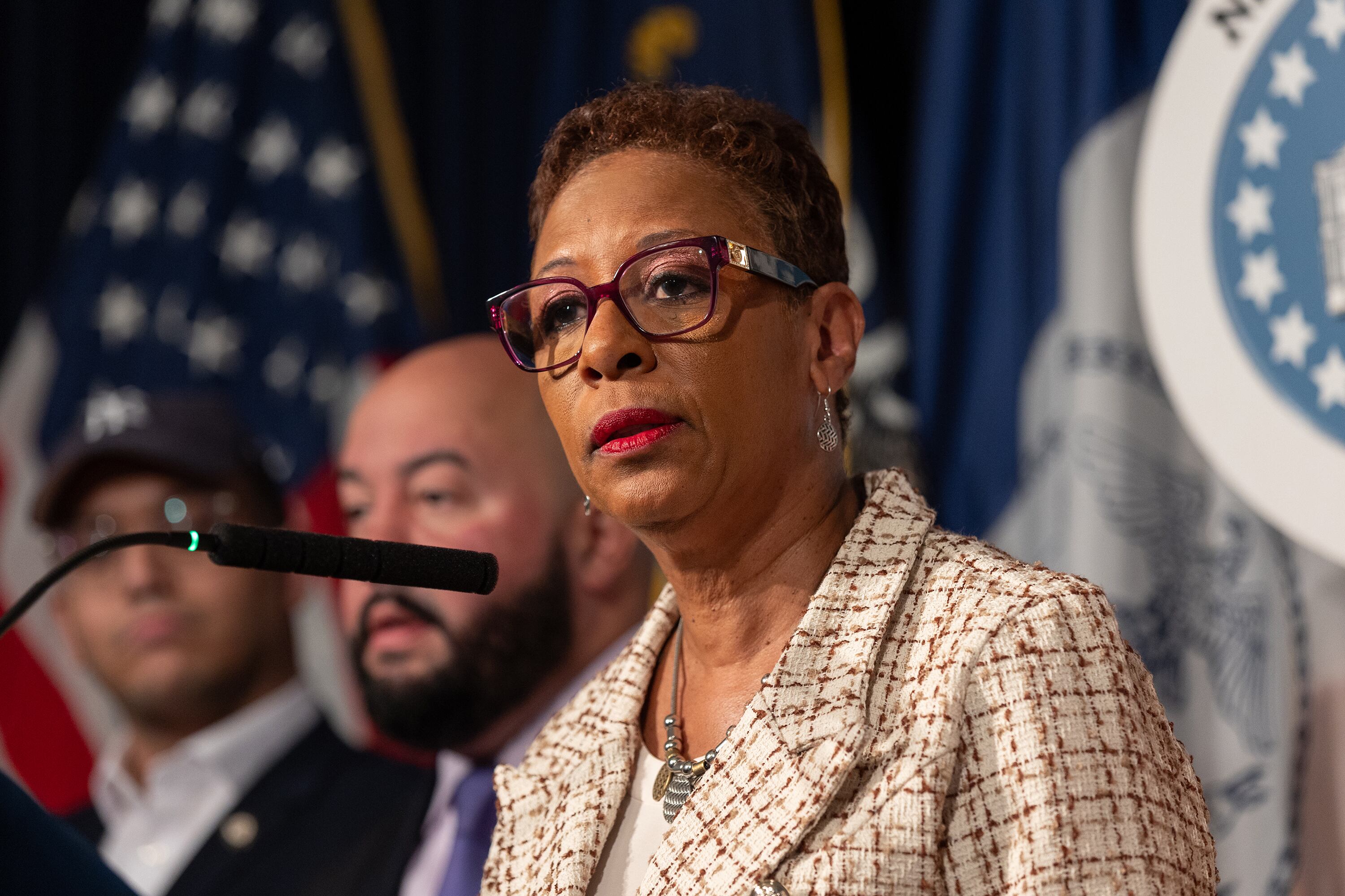 A woman with short dark hair and wearing a tan suit jacket speaks from a microphone with two people and flags in the background.