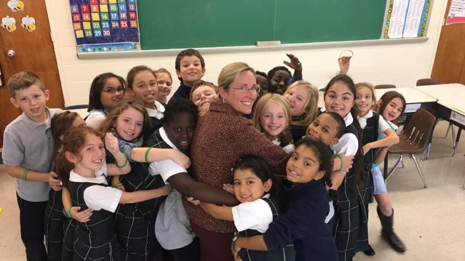 Twenty elementary school students surround Sandy Hook mom Scarlett Lewis and hug her in front of a chalkboard in a classroom.