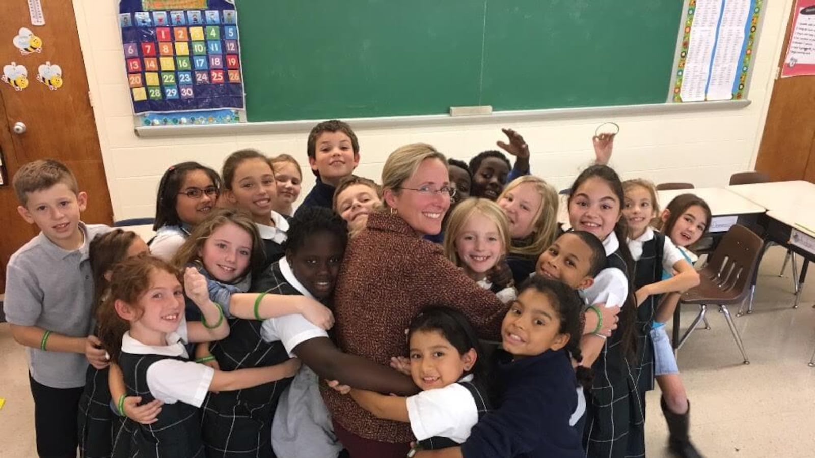 Twenty elementary school students surround Sandy Hook mom Scarlett Lewis and hug her in front of a chalkboard in a classroom. 