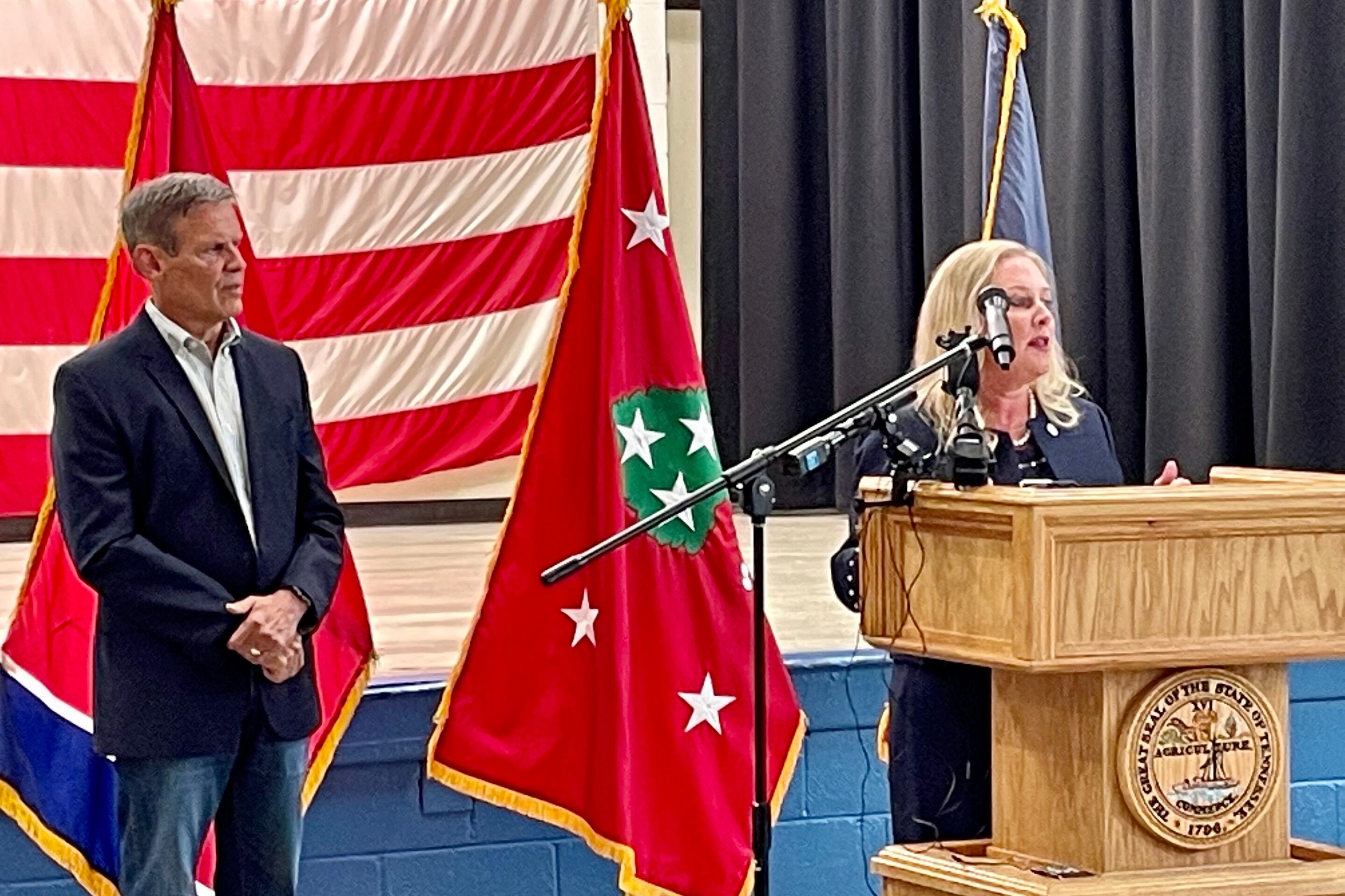 Gov. Bill Lee listens at Health Commissioner Dr. Lisa Piercey talks at a wooden podium.