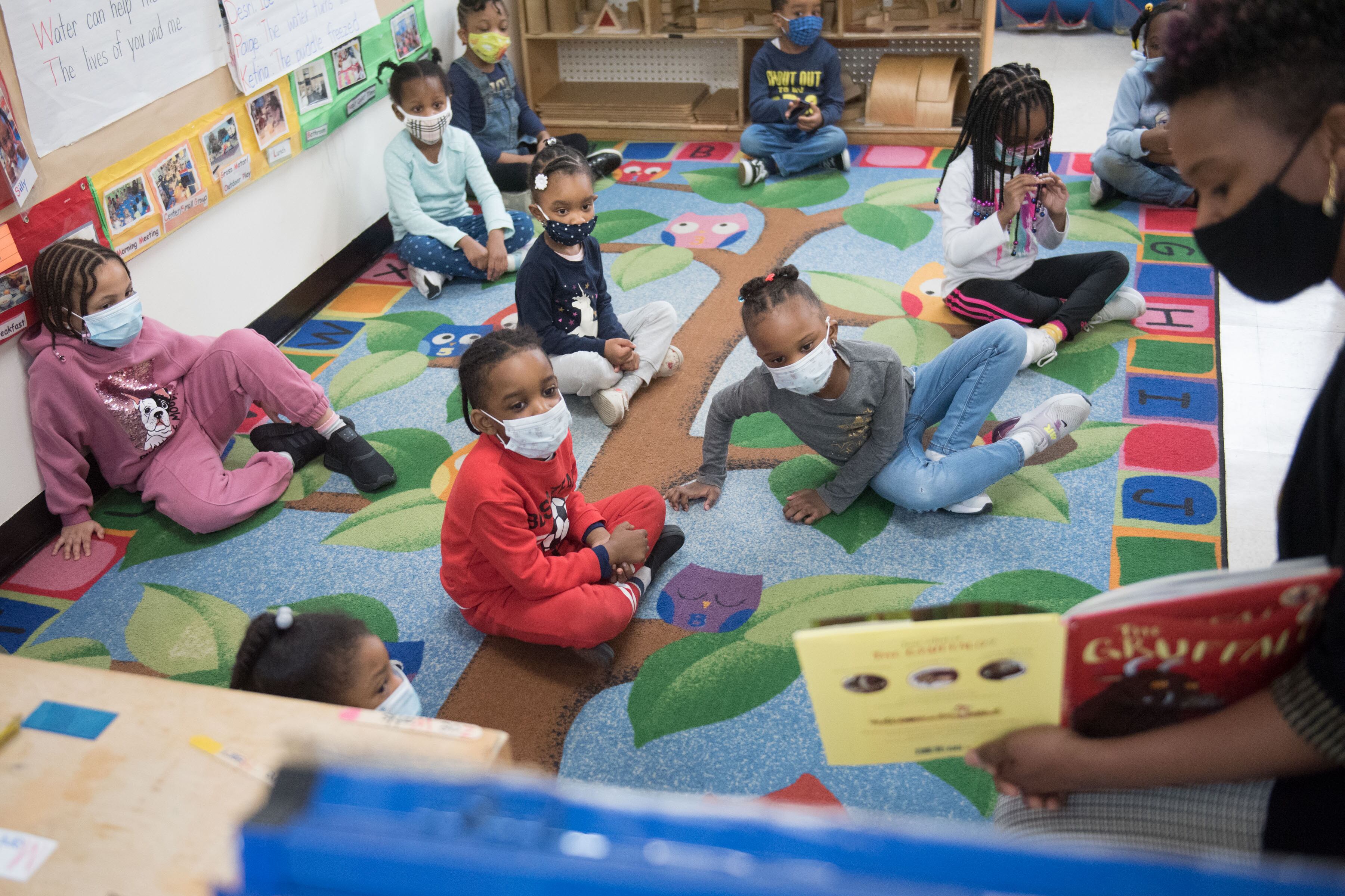 Preschool-age children sit on a colorful rug on the floor as a teacher in a mask reads to them from a yellow and red book.