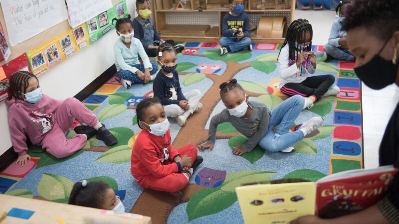 Preschool-age children sit on a colorful rug on the floor as a teacher in a mask reads to them from a yellow and red book.