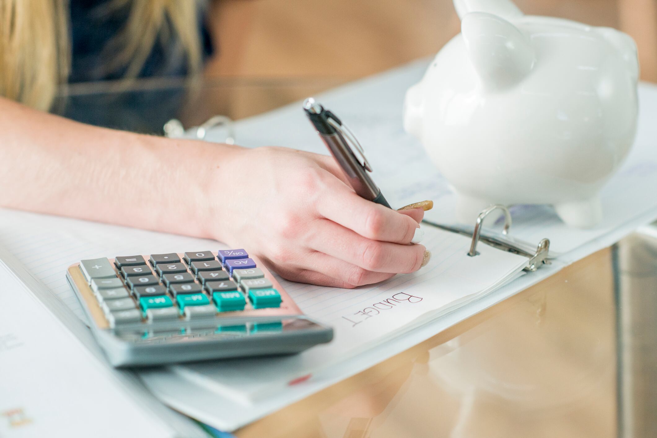 A girl writes in a notebook, with a calculator and white piggy bank sitting on the desk.