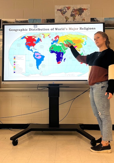 A woman wearing a sweater and jeans points to a projector showing a world map.