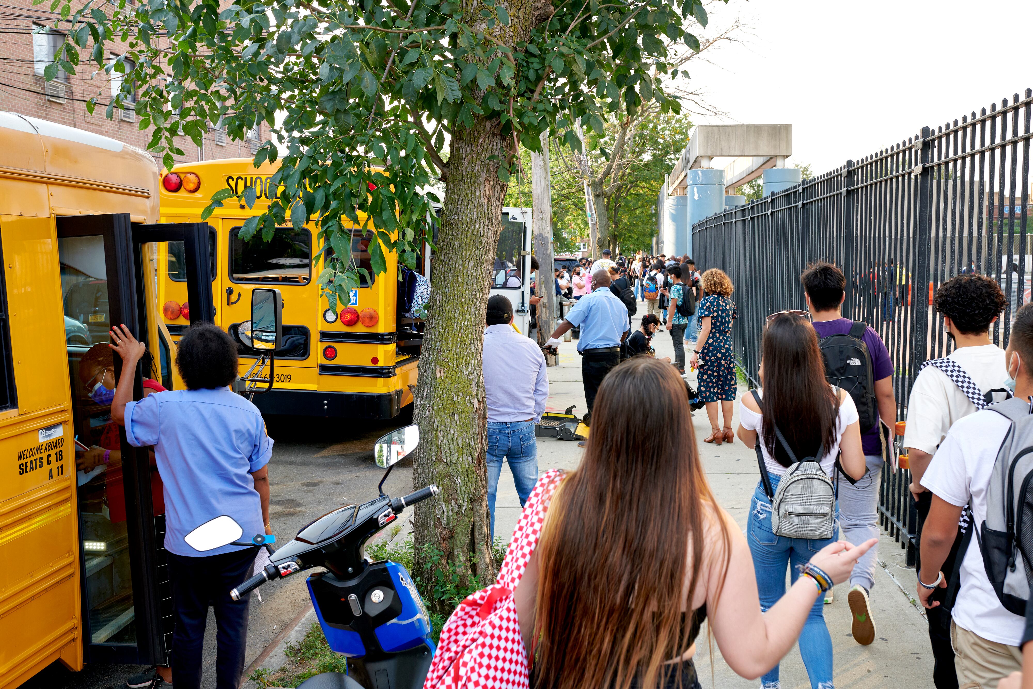 A group of students wearing backpacks flanked by a fence and yellow school buses walk toward a school.