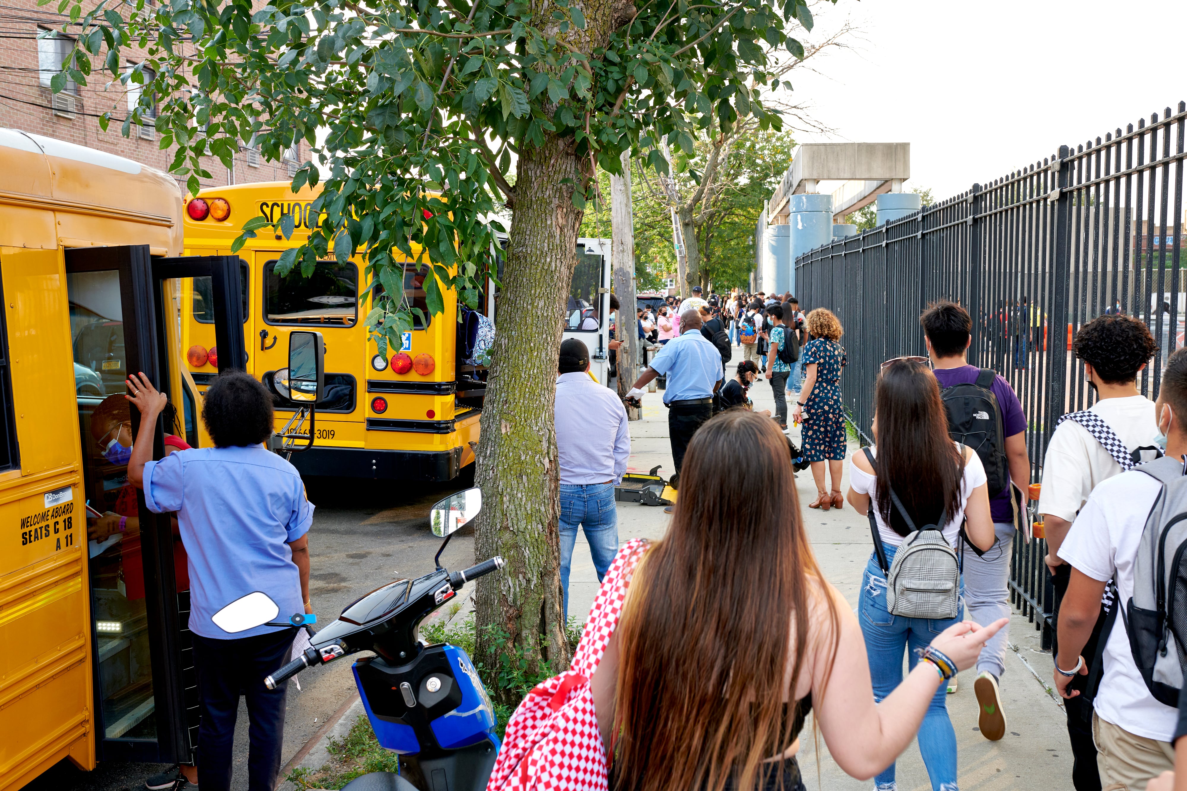 A group of students wearing backpacks flanked by a fence and yellow school buses walk toward a school.