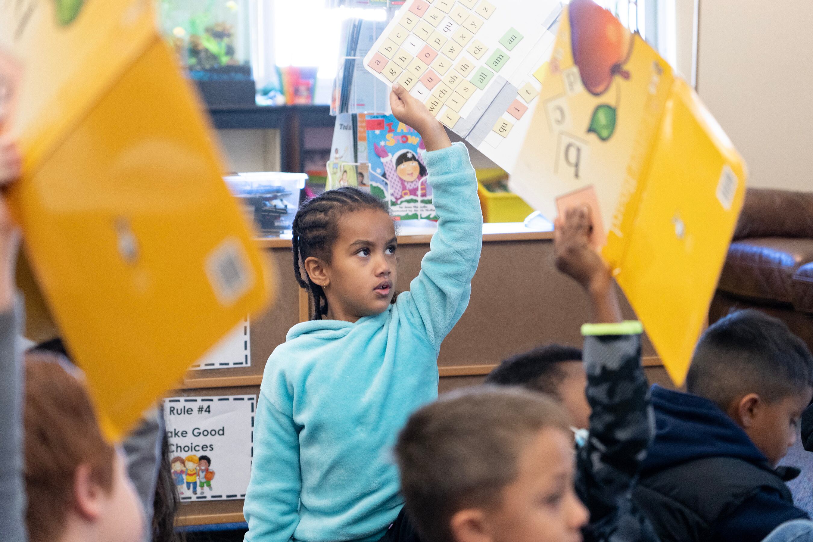 A young girl in a blue sweatshirt, her hair in cornrows, holds up a poster showing her reading lesson.