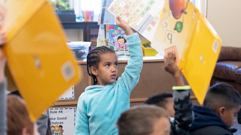 A young girl in a blue sweatshirt, her hair in cornrows, holds up a poster showing her reading lesson.