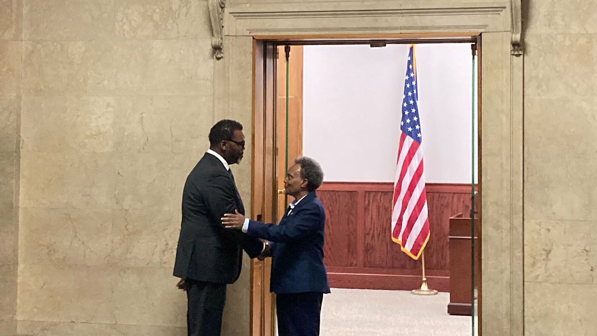 Chicago Mayor-elect Brandon Johnson shakes hands with current Mayor Lori Lightfoot. They are both wearing dark suits and standing in front of a doorway. A U.S. flag is in the background.
