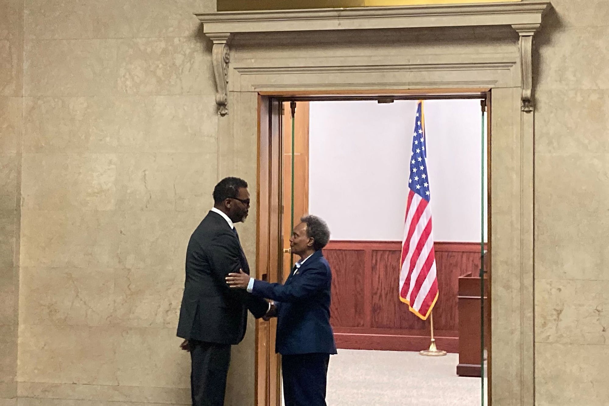 Chicago Mayor-elect Brandon Johnson shakes hands with current Mayor Lori Lightfoot. They are both wearing dark suits and standing in front of a doorway. A U.S. flag is in the background.