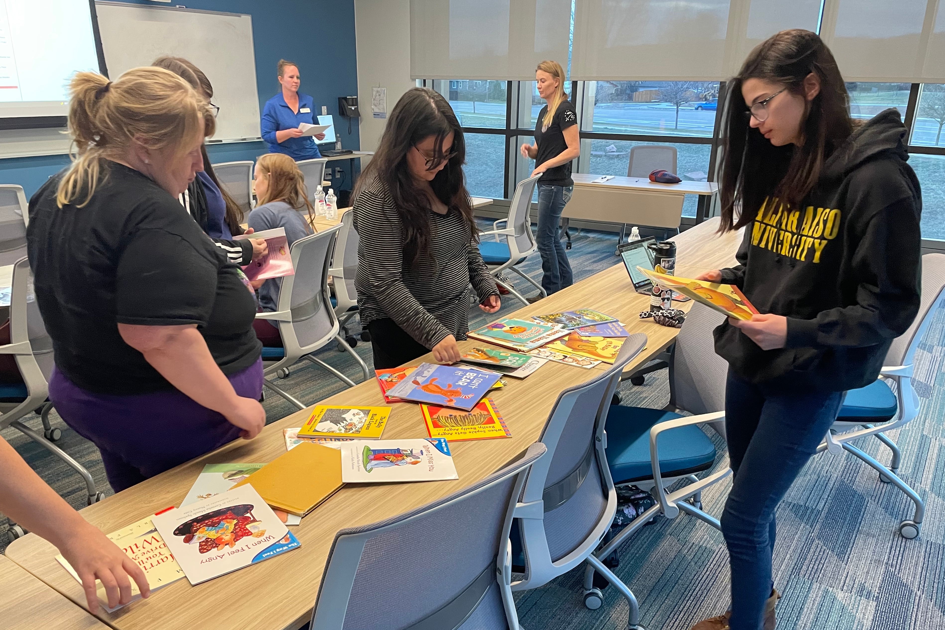 Three students look at children’s books spread out on a table during an evening class.
