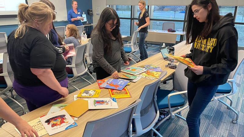 Three students look at children’s books spread out on a table during an evening class.