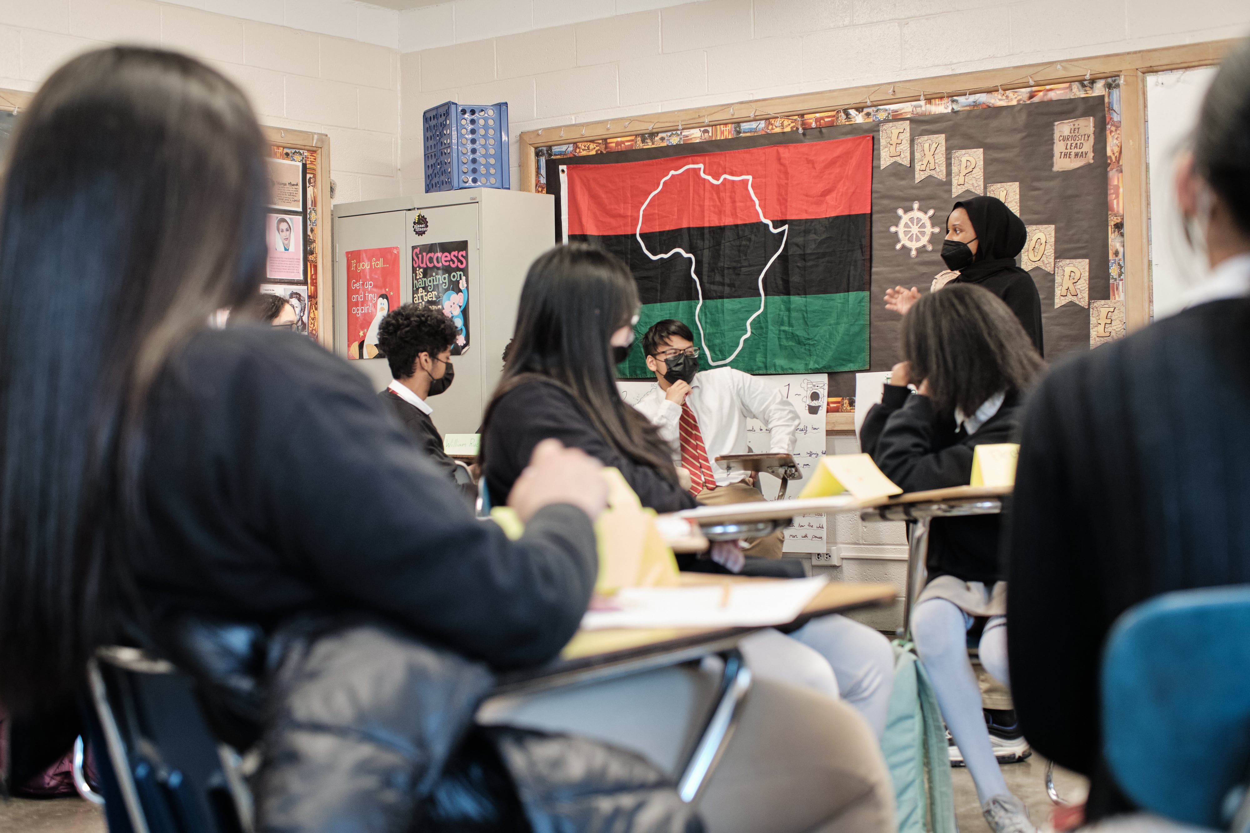 A teacher leads a Black History lecture with her high school class. The students and teacher are wearing masks. There is a large flag with red, black, and green bars and a white outline of Africa behind her.
