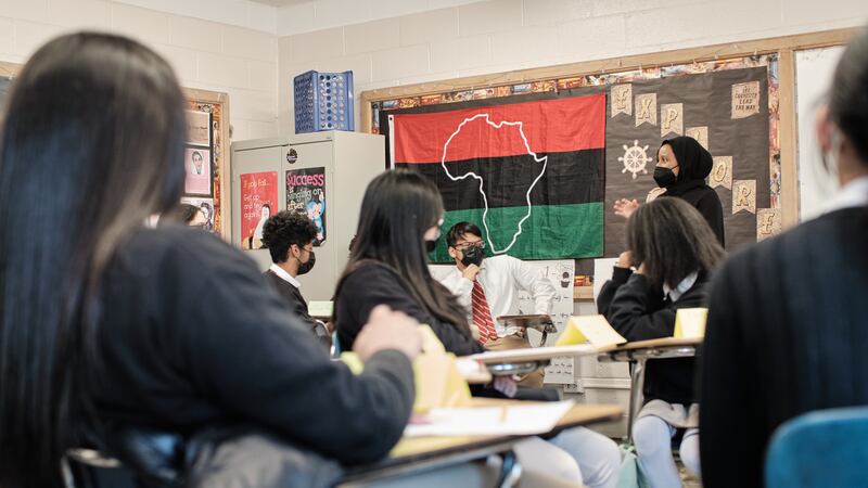 A teacher leads a Black History lecture with her high school class. The students and teacher are wearing masks. There is a large flag with red, black, and green bars and a white outline of Africa behind her.