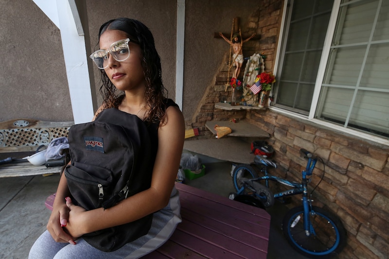 A teenage girl sits on her front porch holding a backpack on her lap. She looks ready for school. In the background, you can see a child's bicycle and a religious shrine.
