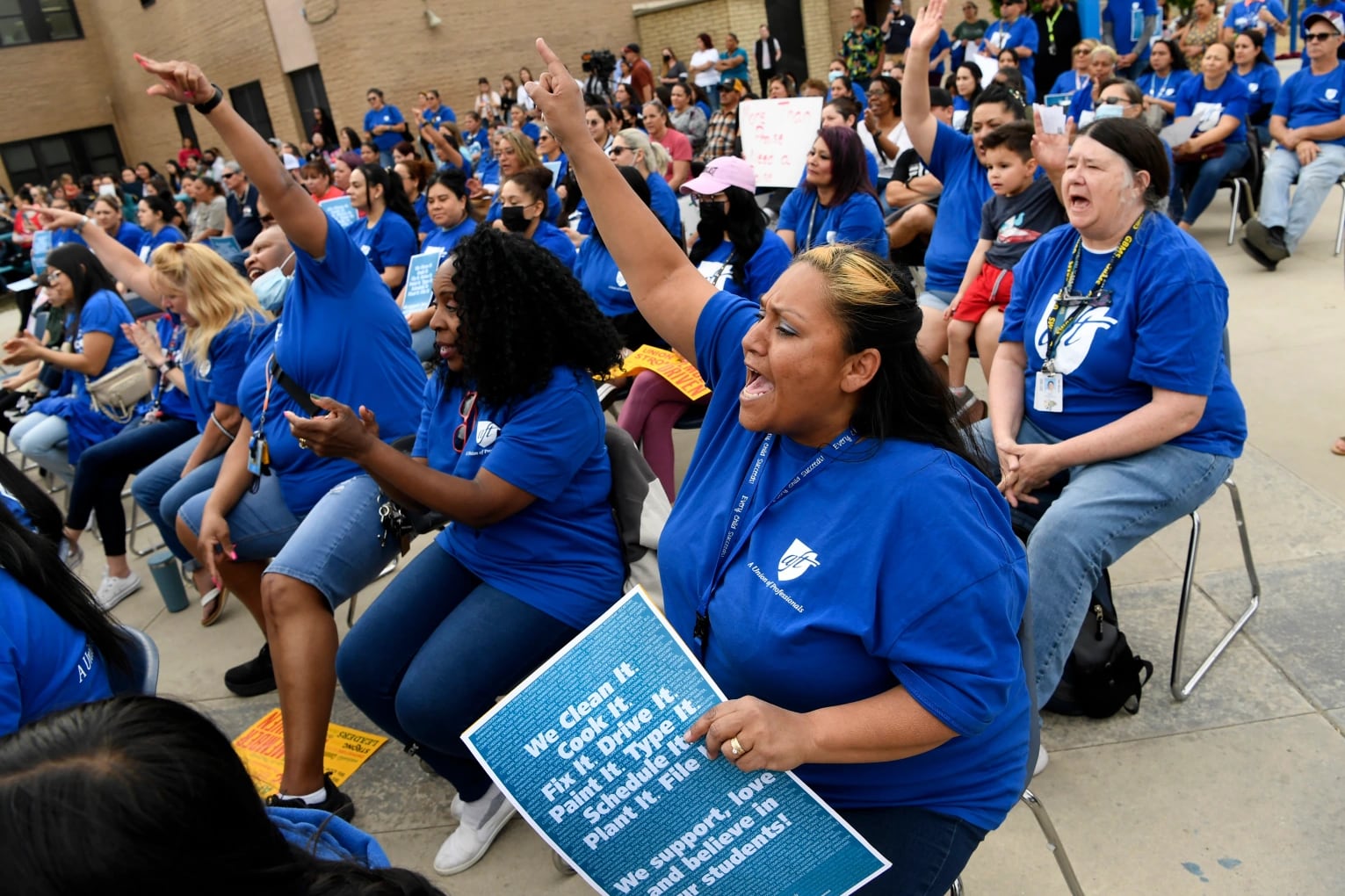 Women wearing matching blue T-shirts seated in chairs shout and cheer with their hands in the air during a rally to increase the minimum wage in Denver Public Schools.