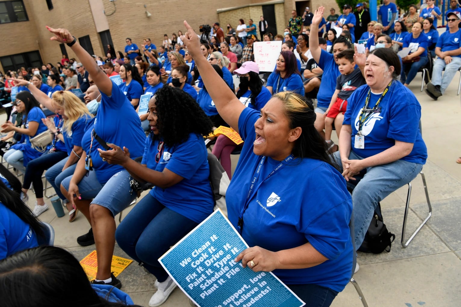 Women wearing matching blue T-shirts seated in chairs shout and cheer with their hands in the air during a rally to increase the minimum wage in Denver Public Schools.