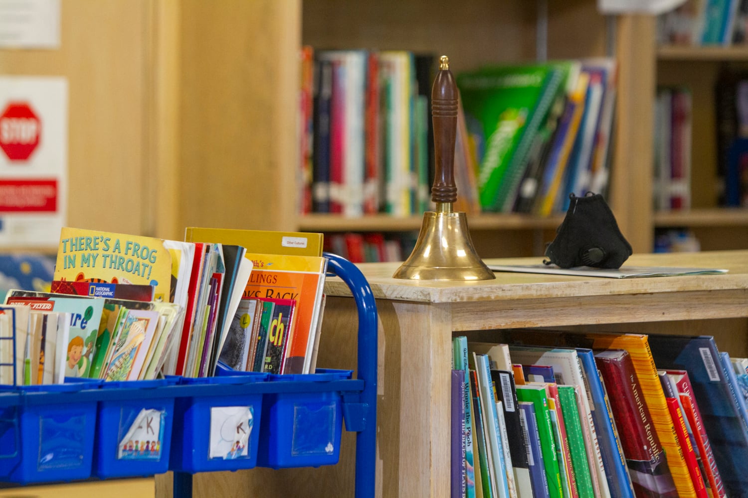A blue book cart with children’s books sits next to full bookshelf with a bell and mask set on top.