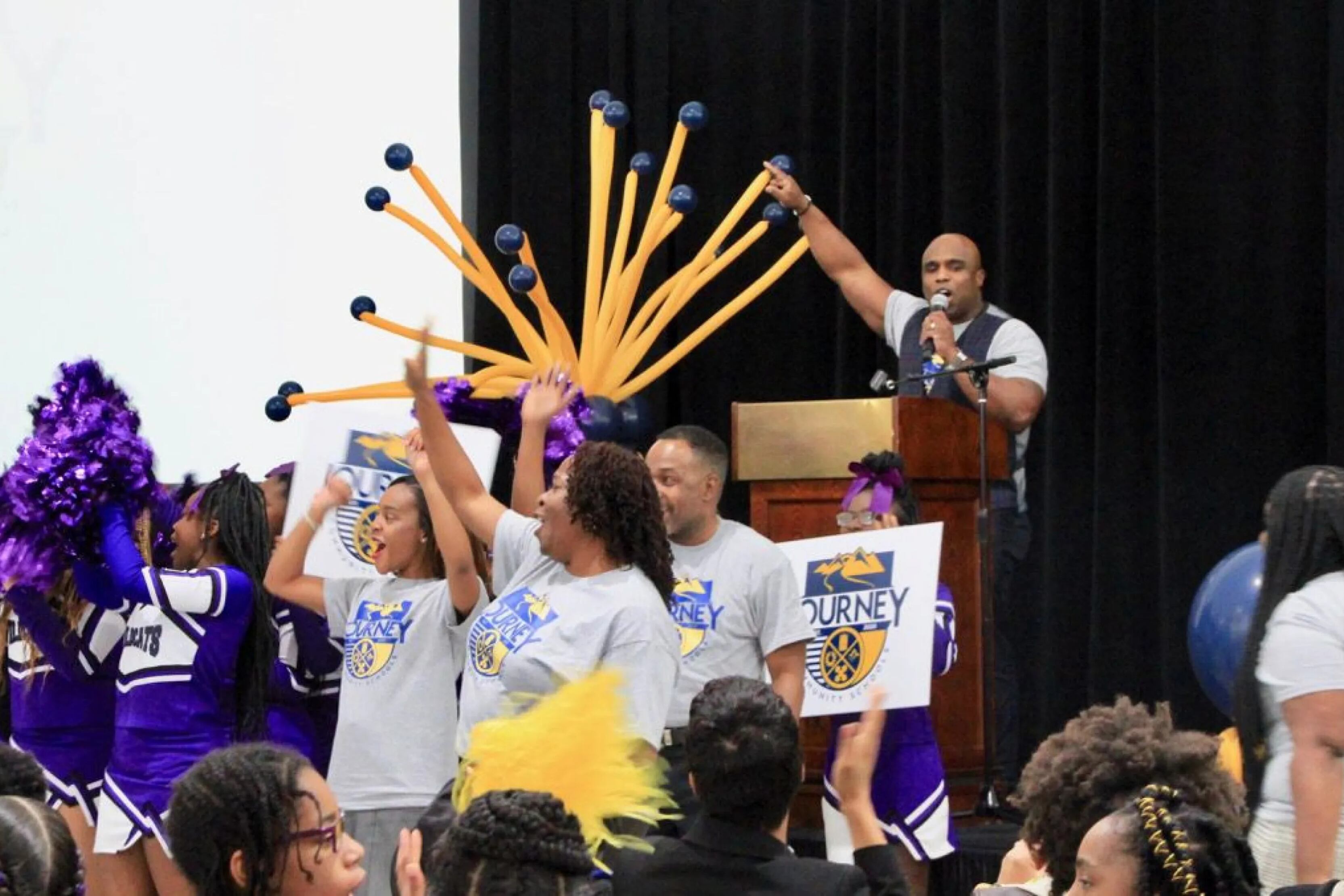 A man behind a podium with arm raised speaks into a microphone on a school stage, behind an enthusiastic crowd that includes cheerleaders.