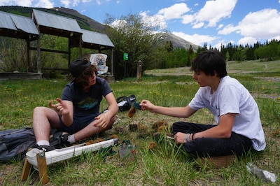 Two teenage boys sit in a green field in the mountains working on some sort of project.