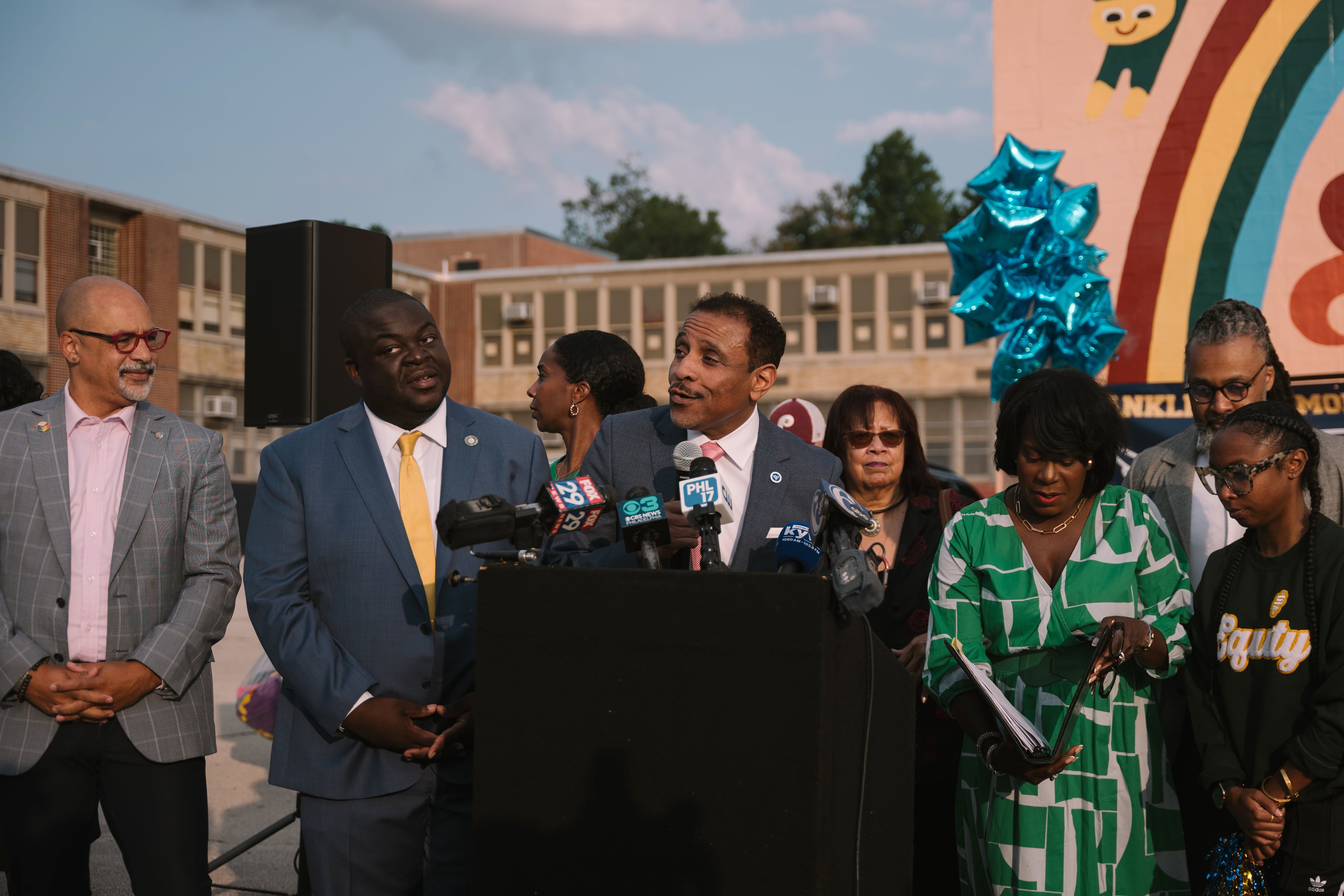 A man in a suit speaks from behind a podium with a group of people in suits standing in the background, all outside.