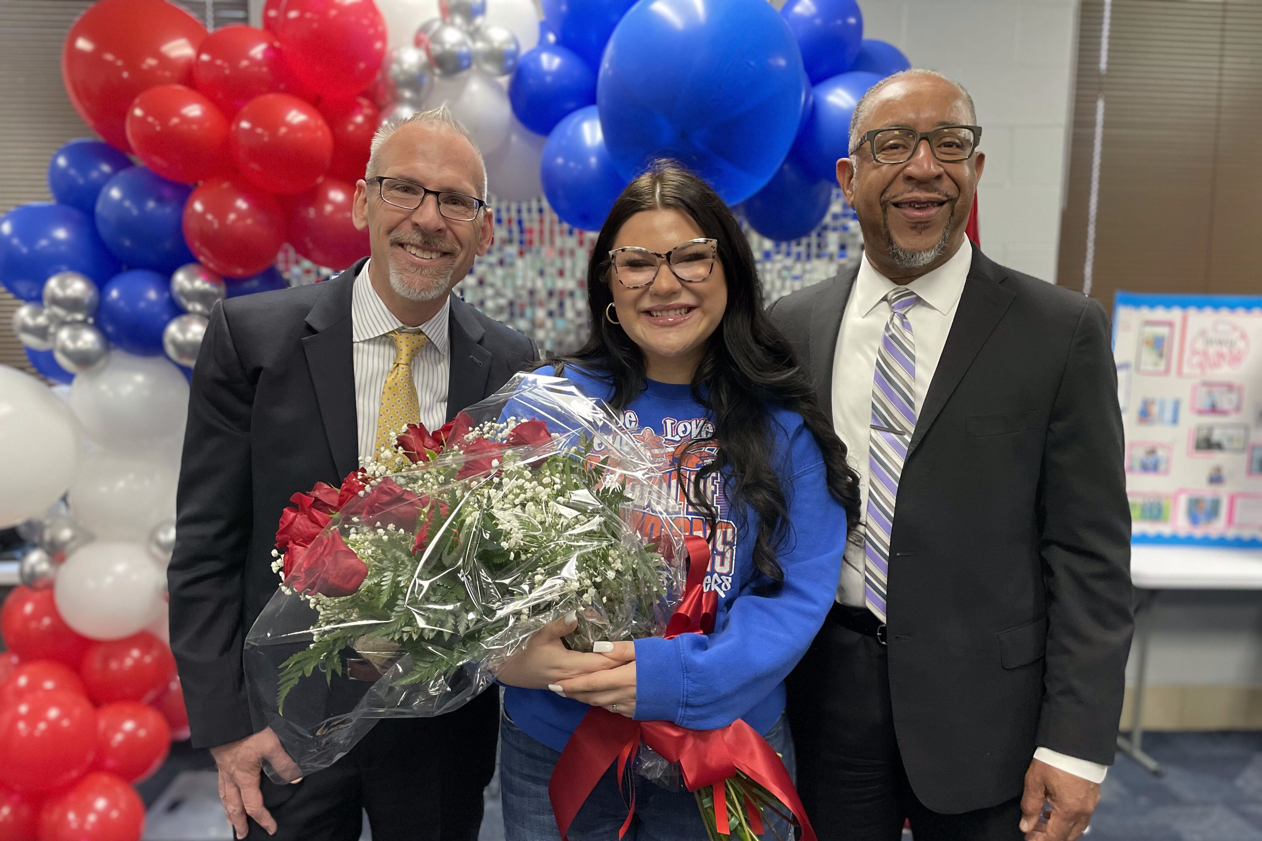 Red, blue, white, and silver balloons are behind State Superintendent Tony Sanders, Briana Morales with a bouquet of flowers in her hands, and Gordon Bush Alternative Center’s principal Darnell Spencer (from left to right) smiling at the camera at a photo.