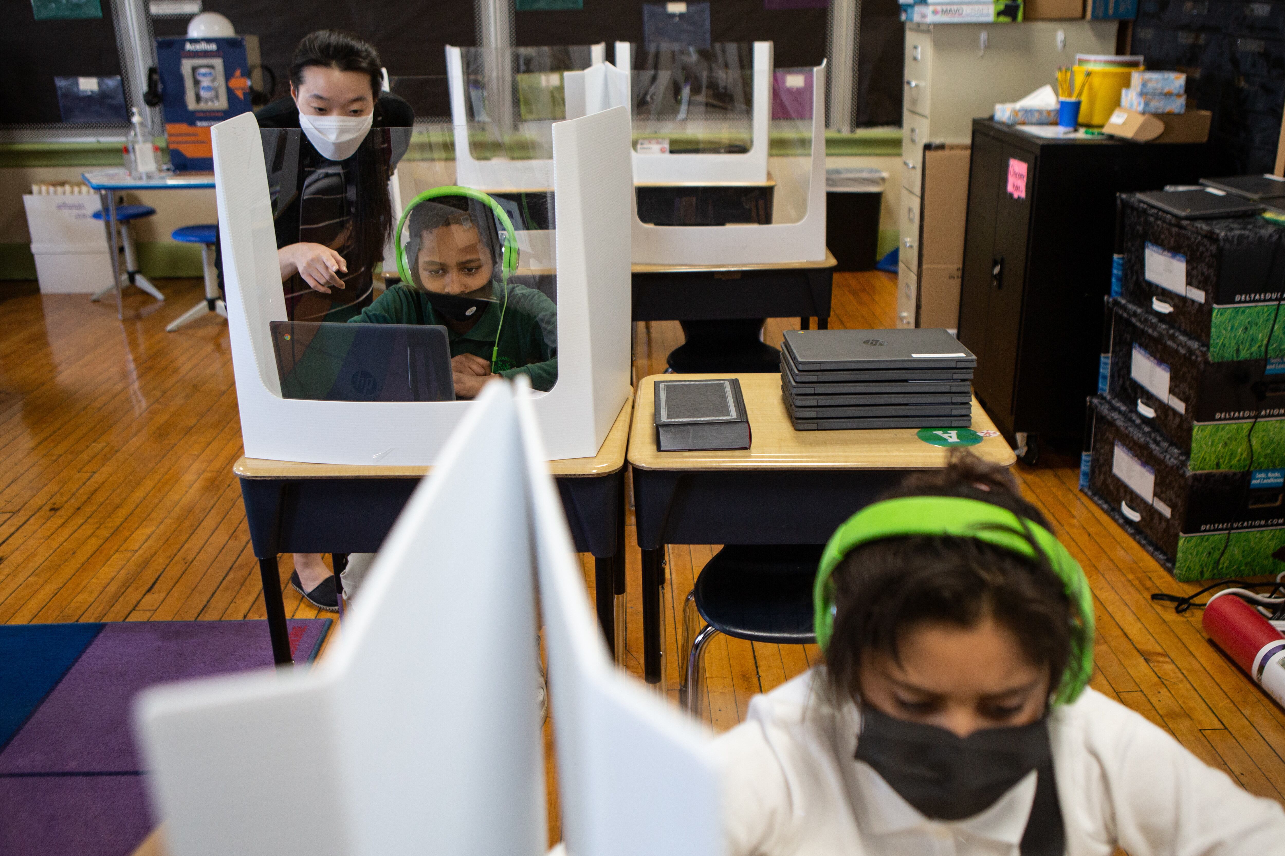 A teacher helps a Roseville fourth grader at his desk while another student wearing green headphones works in the foreground. The classroom’s desks are fitted with partitions aimed at limiting the spread of COVID.
