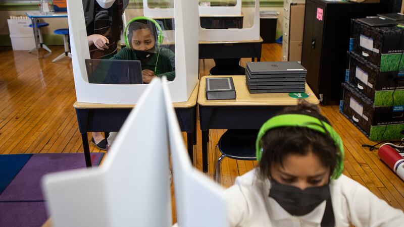 A teacher helps a Roseville fourth grader at his desk while another student wearing green headphones works in the foreground. The classroom’s desks are fitted with partitions aimed at limiting the spread of COVID.