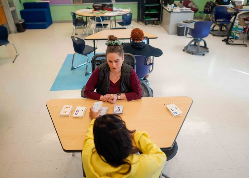 A woman with dark hair in a bun sits at a table with a student in yellow.