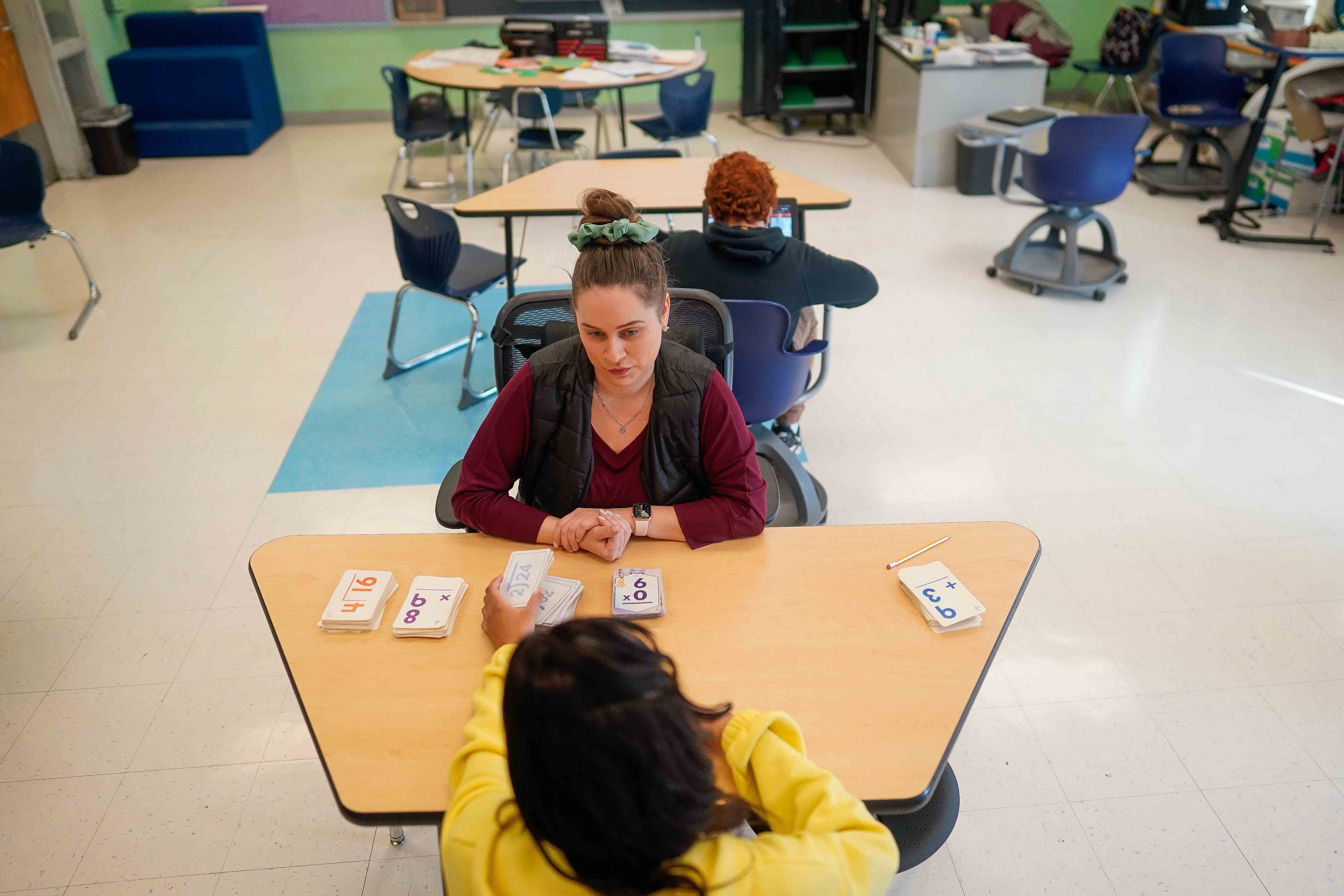 A woman with dark hair in a bun sits at a table with a student in yellow.