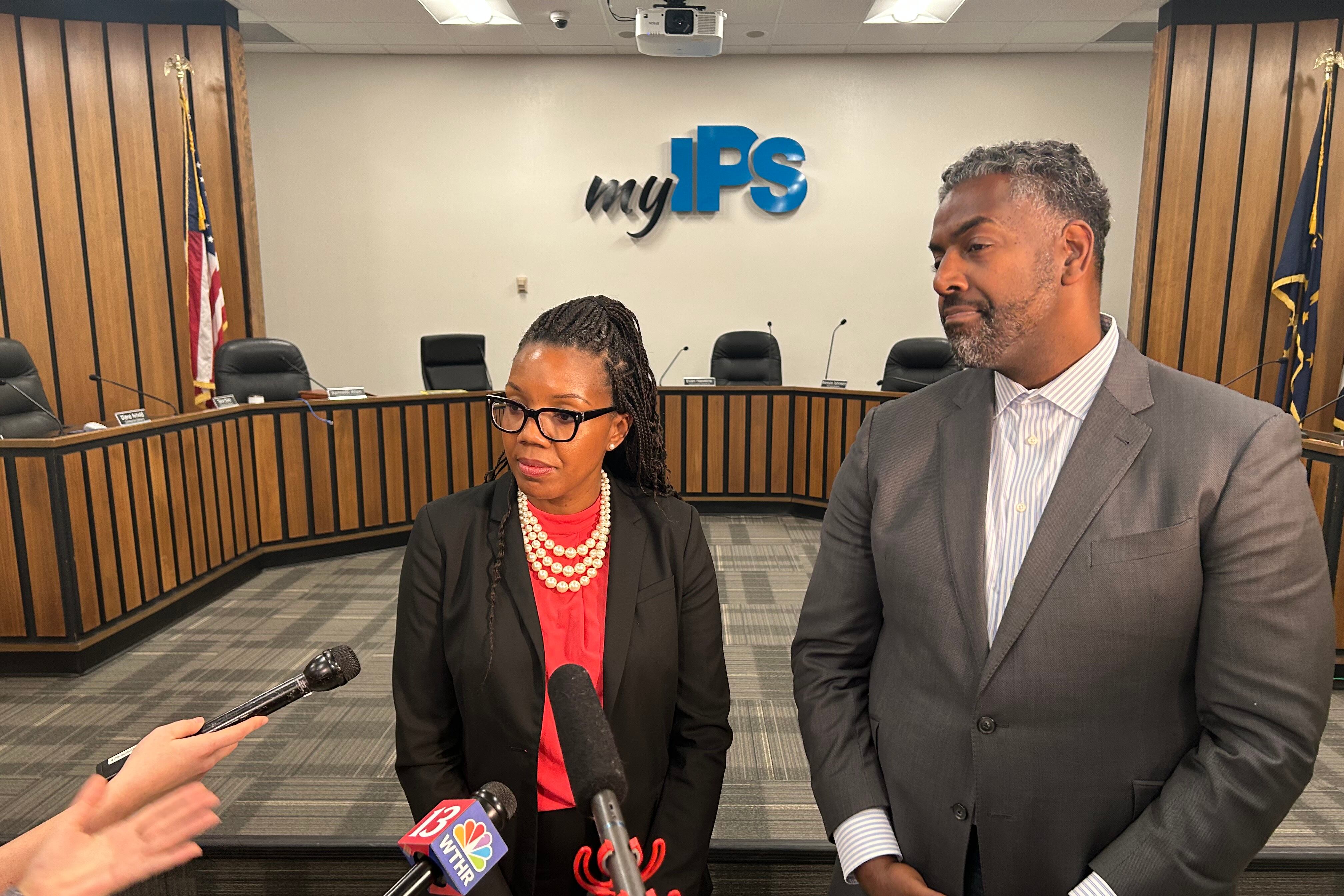 A woman in a black suit jacket and red dress stands in front of a media scrum of microphones and lights next to a man in a suit.