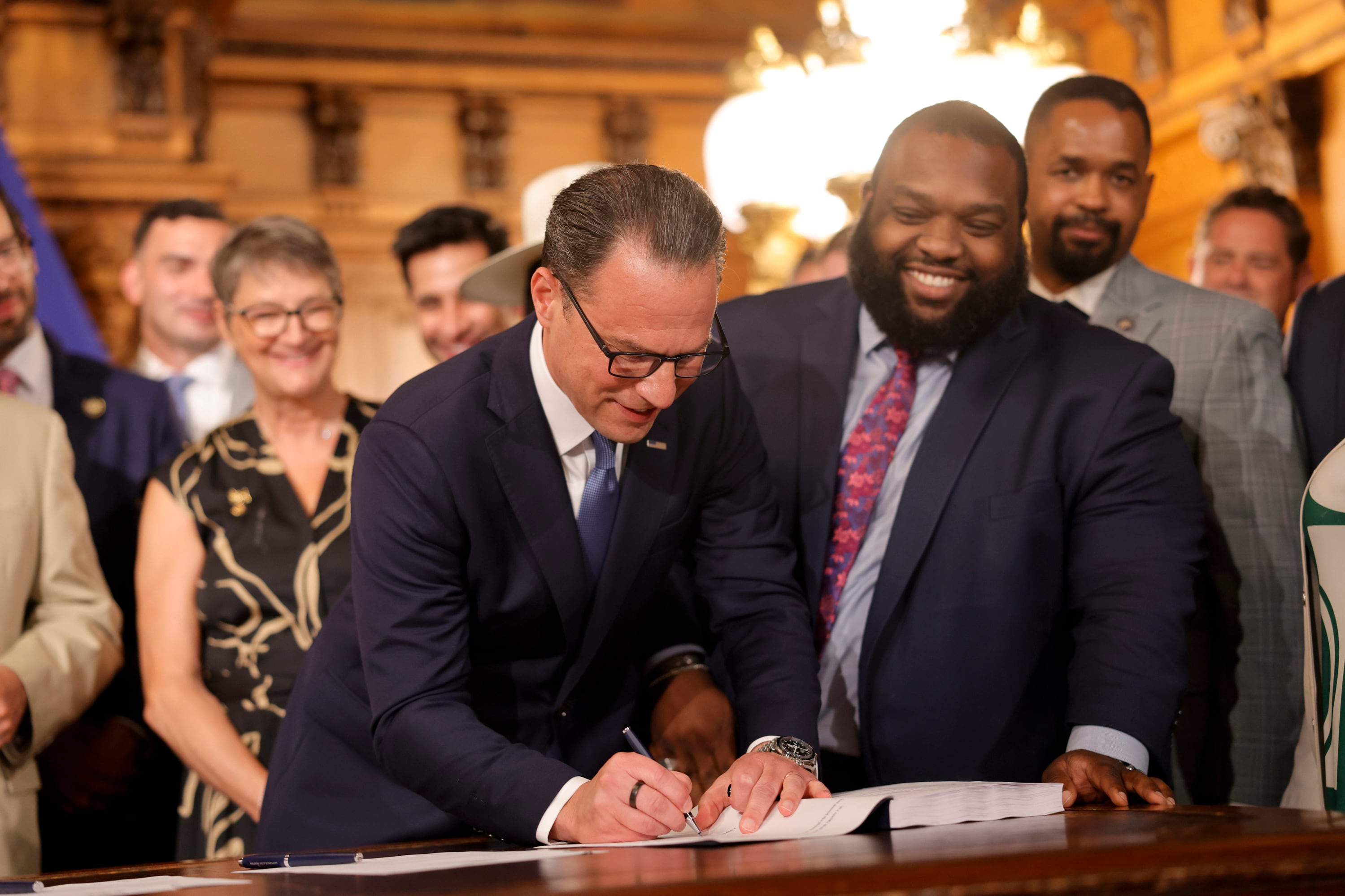 Two men are standing beside a wooden desk; one is signing a document with a pen.
