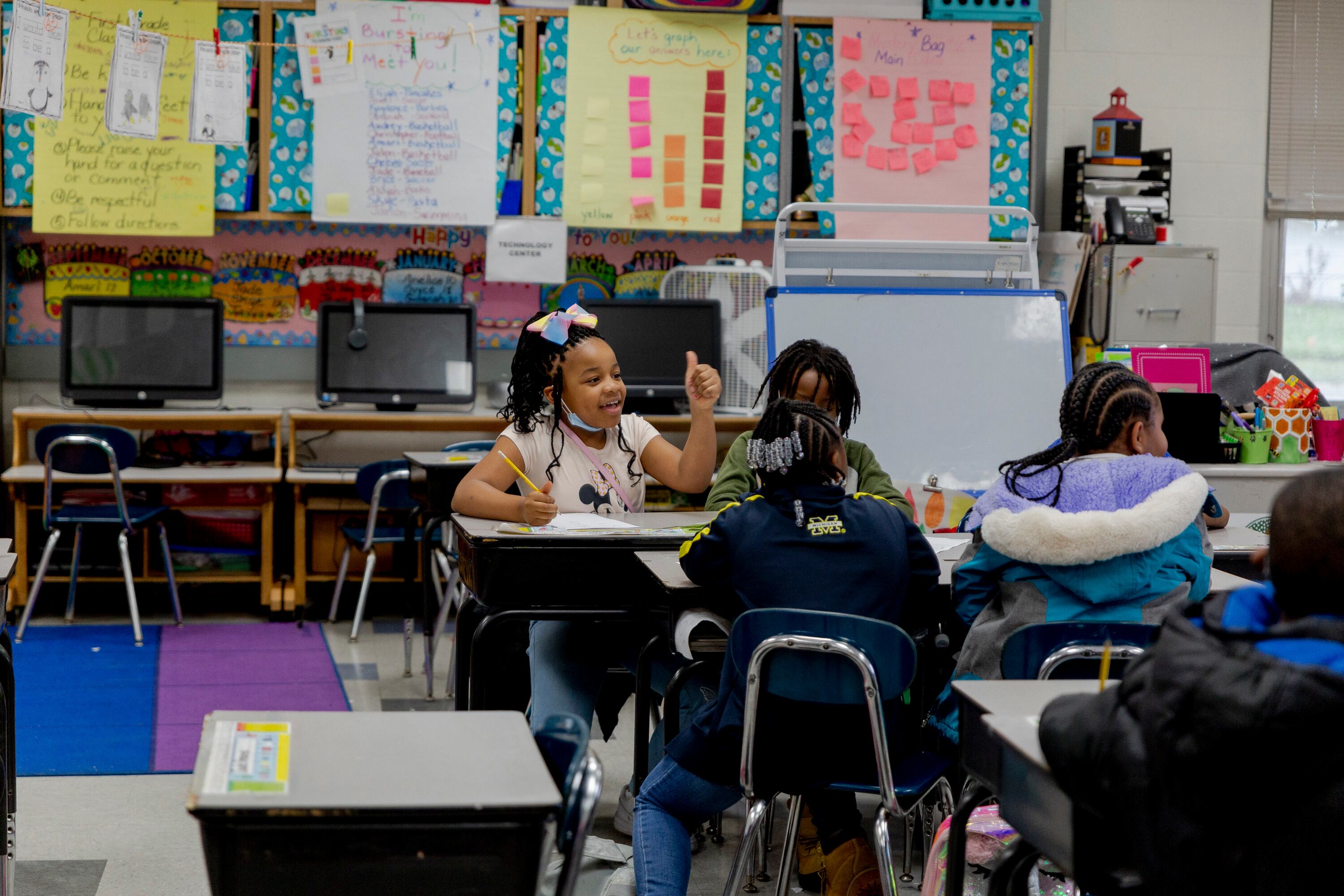 A young girl with a bow in her hair sits at a desk and talks with three other students in a classroom with colorful bulletin boards.