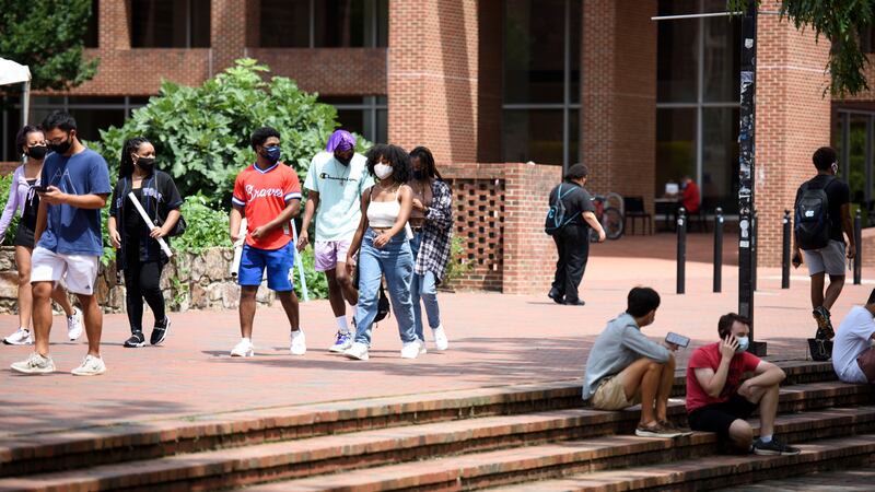 Small groups of students walk on a university campus plaza, with a building in the background. An handful of students sit on the steps of the plaza.