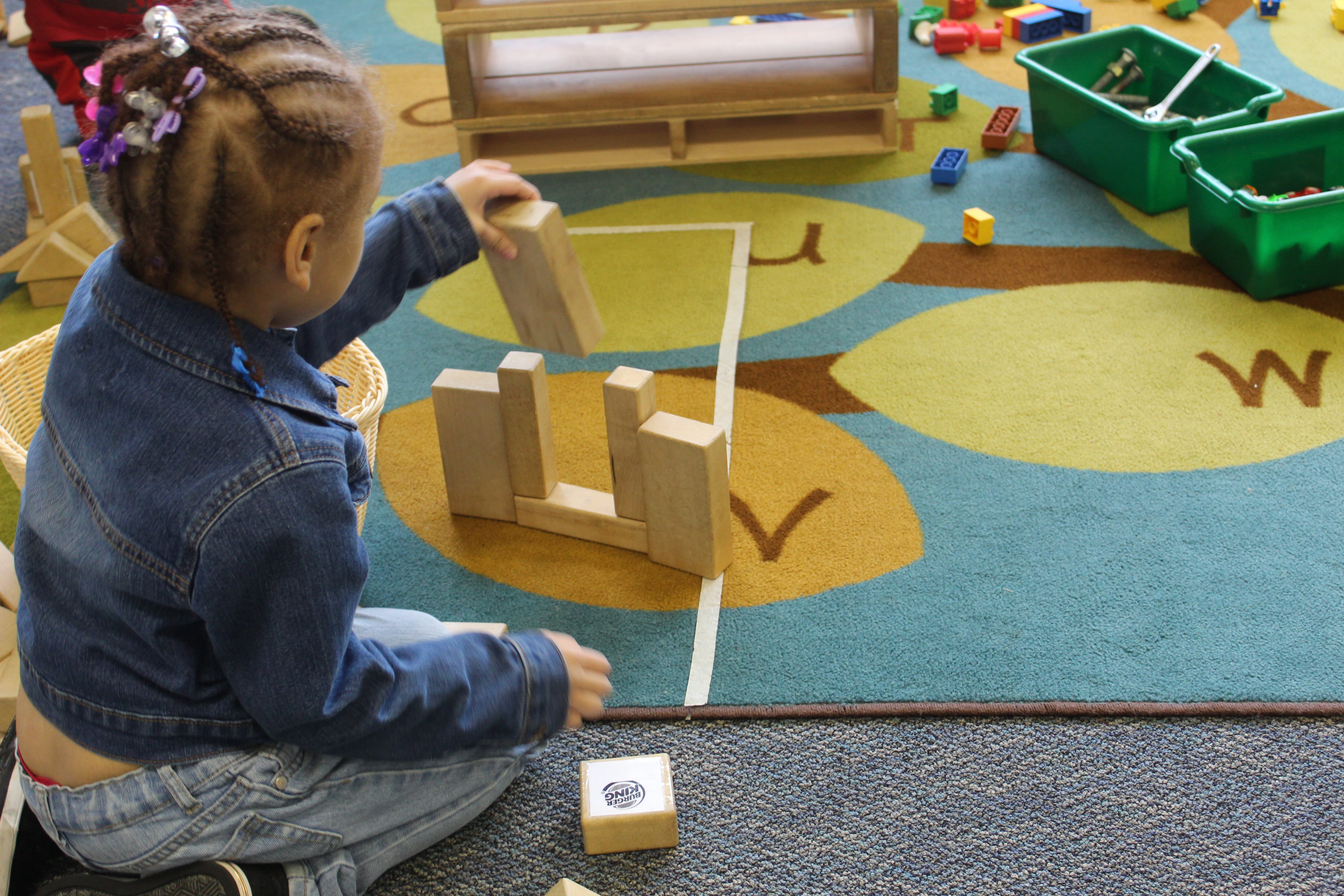 A photograph of a young preschooler playing on a colorful rug with wooden blocks.