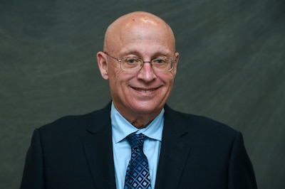 A man wearing glasses and a dark suit poses for a portrait in front of a green background.