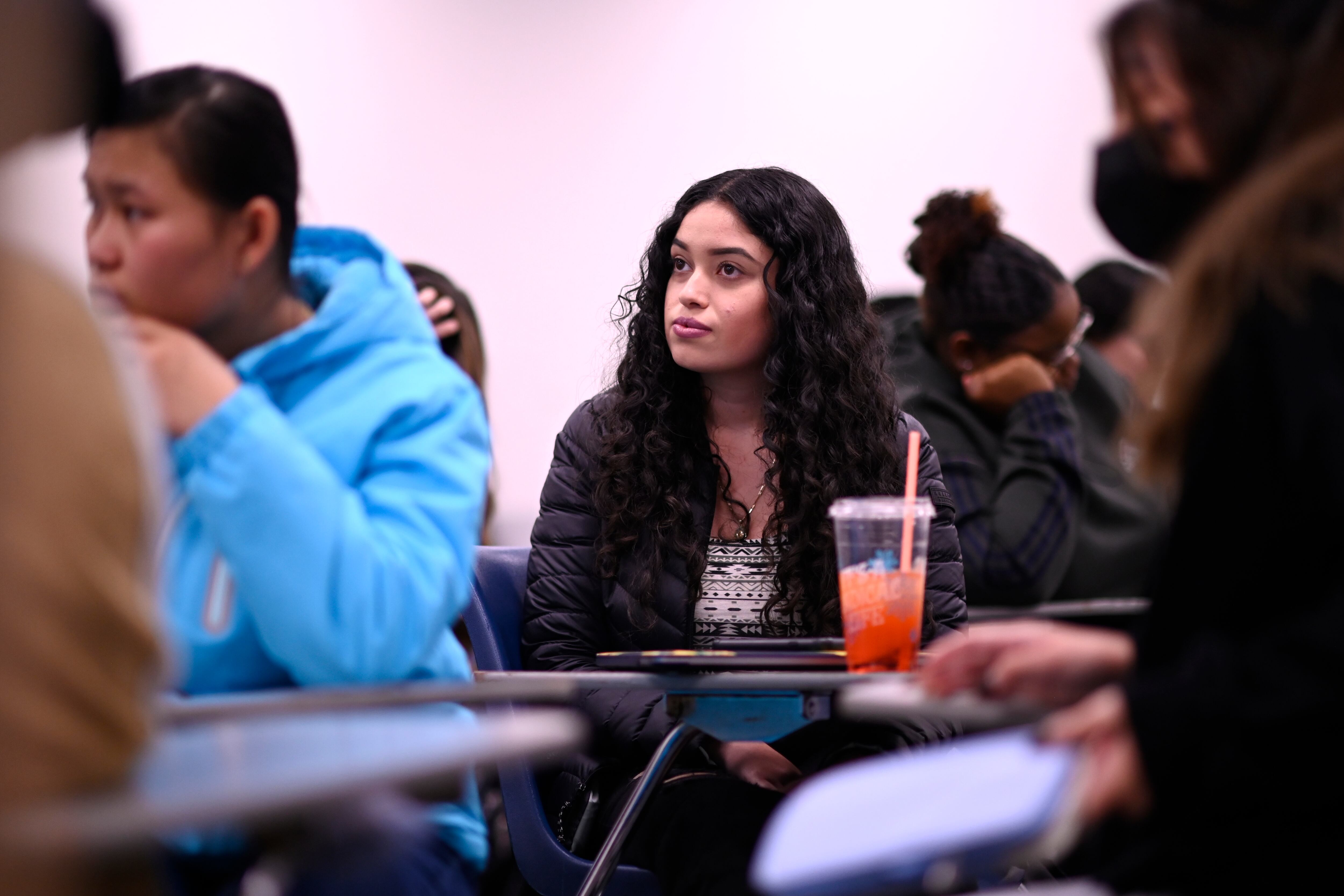 A young woman with long dark hair sits in a crowded college classroom. She looks toward the front of the room. She has a large orange-pink drink on her desk. She’s surrounded by other students.