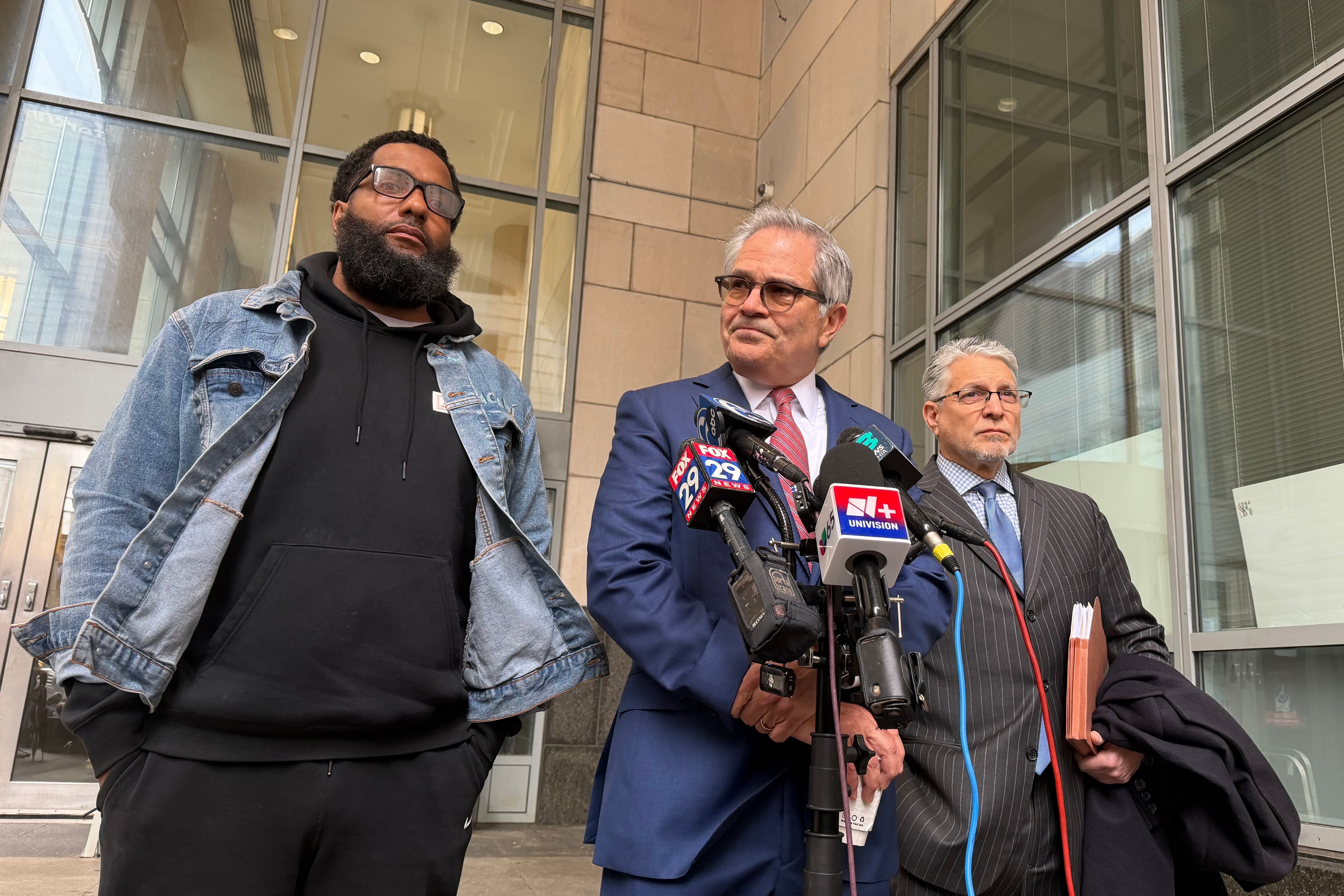 A photograph of one Black man and two white men standing by press microphones outside speaking during a press conference.